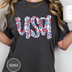May include: A dark gray t-shirt with the word "USA" printed in white outlined in black. The letters are filled with a red, white, and blue floral pattern.