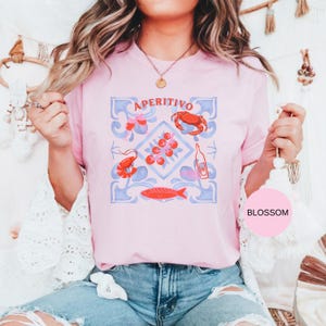Op de afbeelding: Lichtroze T-shirt met het woord "APERITIVO" en een grafisch ontwerp met een krab, garnalen, vis, tomaten en een fles. Het ontwerp is in rood, blauw en wit. Een casual kledingstuk.