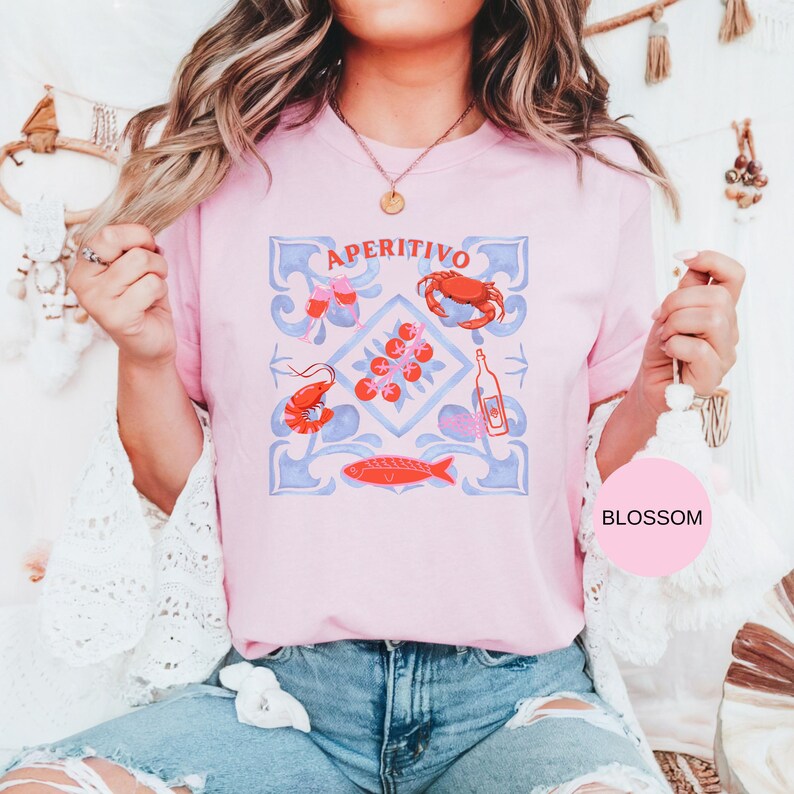 Op de afbeelding: Lichtroze T-shirt met het woord "APERITIVO" en een grafisch ontwerp met een krab, garnalen, vis, tomaten en een fles. Het ontwerp is in rood, blauw en wit. Een casual kledingstuk.
