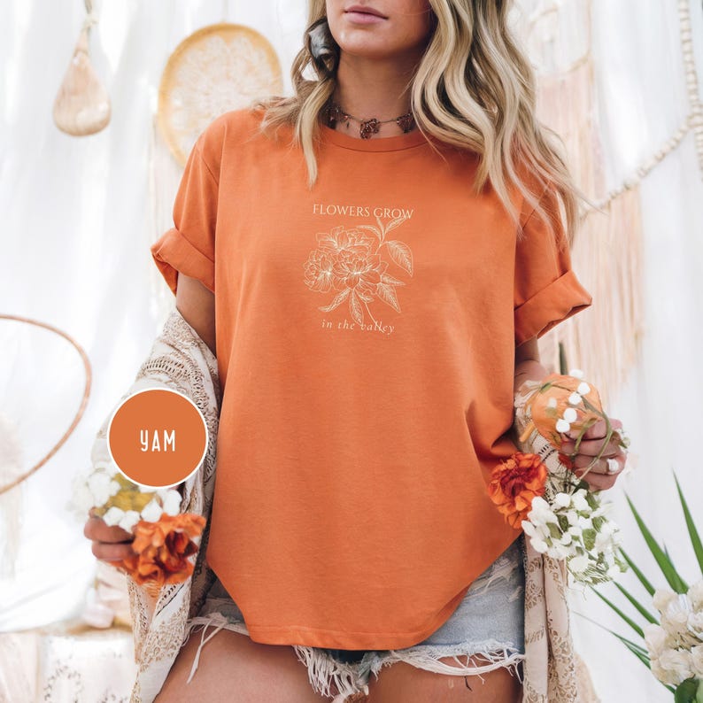 May include: Orange t-shirt with rolled sleeves featuring the text "FLOWERS GROW in the valley" and a floral design. The shirt is a solid orange color. The image also includes a small round sticker with the letters "YAM".