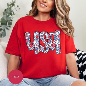 May include: A red t-shirt with a white and blue floral design that spells out "USA".