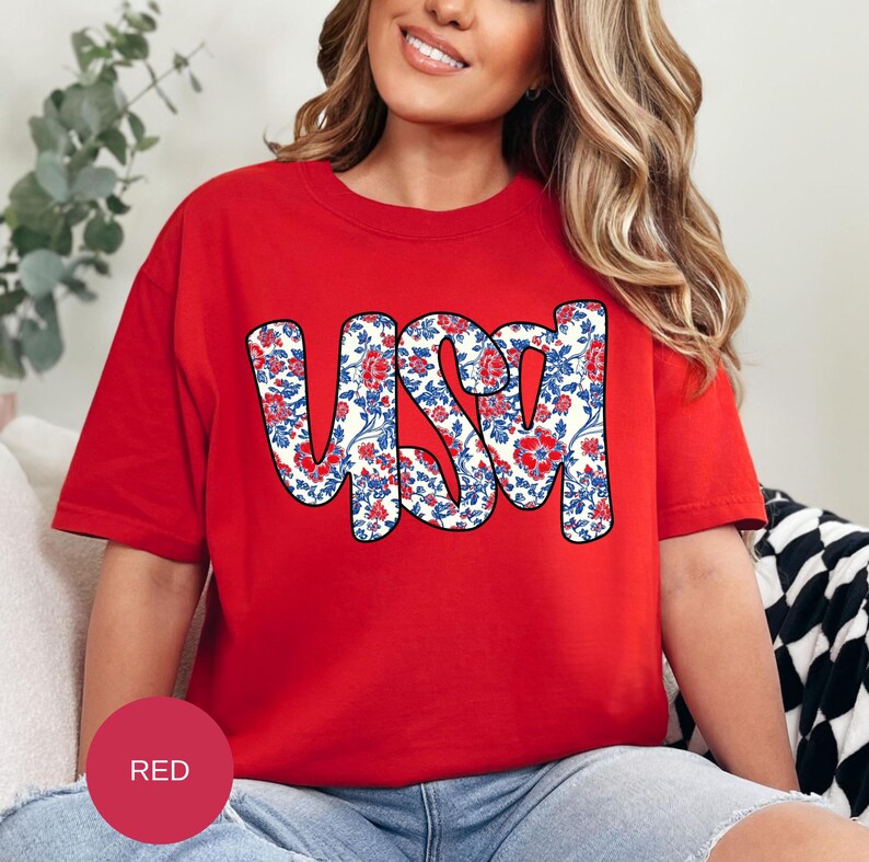 May include: A red t-shirt with a white and blue floral design that spells out "USA".