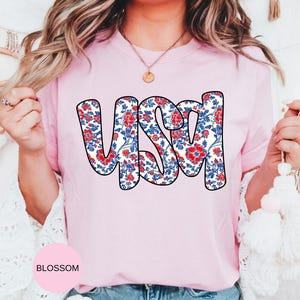May include: A pink t-shirt with a floral pattern spelling out "USA" in a bold, white outline.