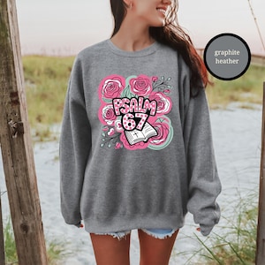 May include: A graphite heather sweatshirt featuring a floral design with pink roses and the text "PSALM 67". The design includes an open book with a cross. The sweatshirt is a comfortable, casual style.