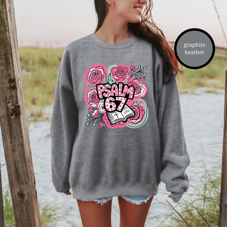 May include: A graphite heather sweatshirt featuring a floral design with pink roses and the text "PSALM 67". The design includes an open book with a cross. The sweatshirt is a comfortable, casual style.