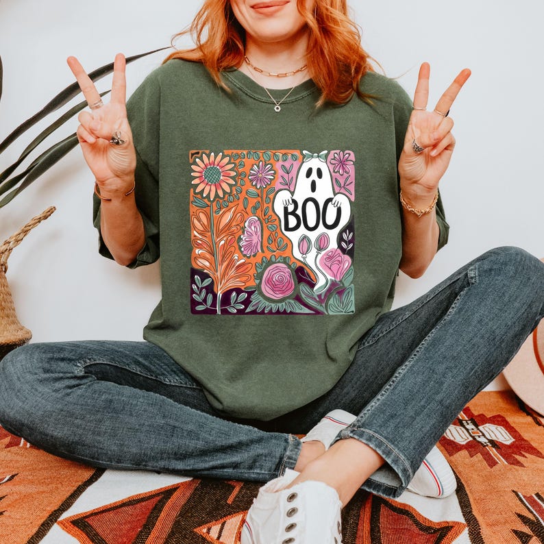 May include: Dark green sweatshirt featuring a colorful floral and ghost graphic. The graphic includes a white ghost with the word "BOO" and various flowers in shades of orange, pink, and purple. The sweatshirt is a relaxed fit.