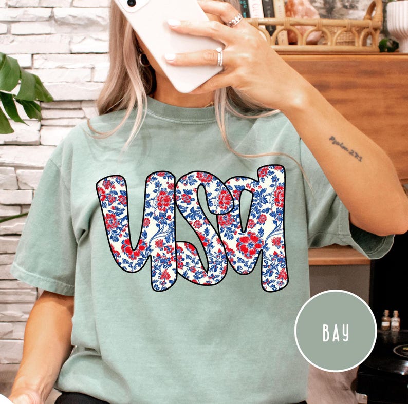 May include: A light green t-shirt with a floral pattern spelling out "USA" in bold black letters. The floral pattern is red, white, and blue.