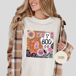 May include: Ivory-colored t-shirt featuring a floral design with a ghost that says "BOO". The design includes orange, pink, and purple flowers. The shirt is a crew neck and has short sleeves. The shirt is made of a soft, comfortable material.