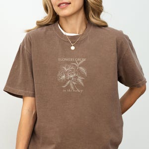 May include: A brown t-shirt with the text "FLOWERS GROW in the valley" and a floral graphic. The shirt has short sleeves and a crew neck. The graphic is in white.