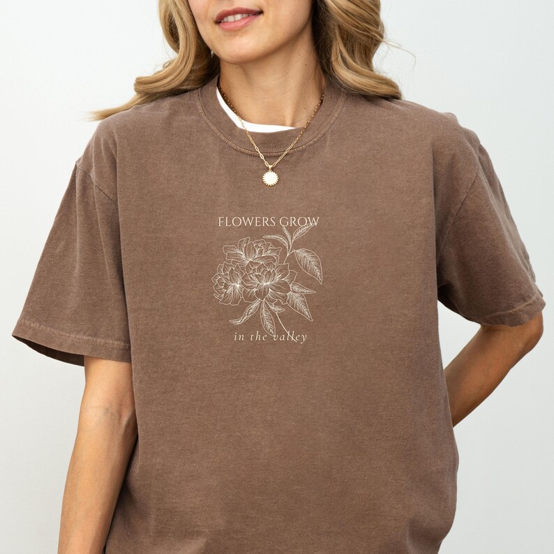 May include: A brown t-shirt with the text "FLOWERS GROW in the valley" and a floral graphic. The shirt has short sleeves and a crew neck. The graphic is in white.