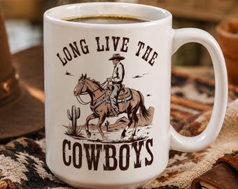 May include: White ceramic mug with a handle, featuring a cowboy riding a horse illustration. The mug has the text "LONG LIVE THE COWBOYS" in a brown font. The mug is filled with a dark liquid, possibly coffee.