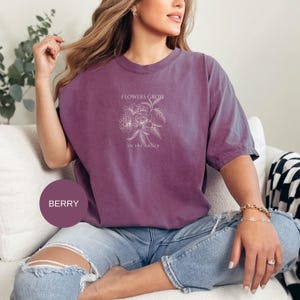 May include: Berry-colored t-shirt with a floral design and the text "FLOWERS GROW in the valley". The shirt is a relaxed fit and is paired with blue jeans. The shirt is a solid color with a crew neck.