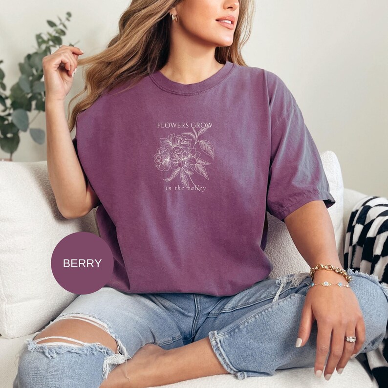 May include: Berry-colored t-shirt with a floral design and the text "FLOWERS GROW in the valley". The shirt is a relaxed fit and is paired with blue jeans. The shirt is a solid color with a crew neck.