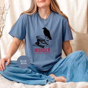 May include: Blue jean t-shirt with a black raven perched atop a stack of books. The word "MURDER" is printed in red, with the phrase "but make it literary" below. The shirt is a solid blue color.