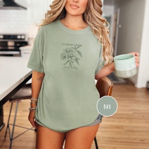 May include: Sage green t-shirt with short sleeves. The shirt features a floral line drawing and the text "FLOWERS GROW in the valley". A round color swatch labeled "BAY" is in the lower right corner.