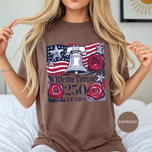 May include: Espresso-coloured t-shirt with a patriotic design. The graphic features the American flag, a silver Liberty Bell, red roses, and the text "We the People 250 Years". The shirt is a comfortable, casual style.