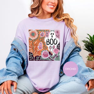May include: A light purple t-shirt with a colorful graphic featuring a white ghost with the word "BOO" and floral elements. The design includes orange, pink, and green flowers. The shirt is part of a Halloween or fall-themed collection.