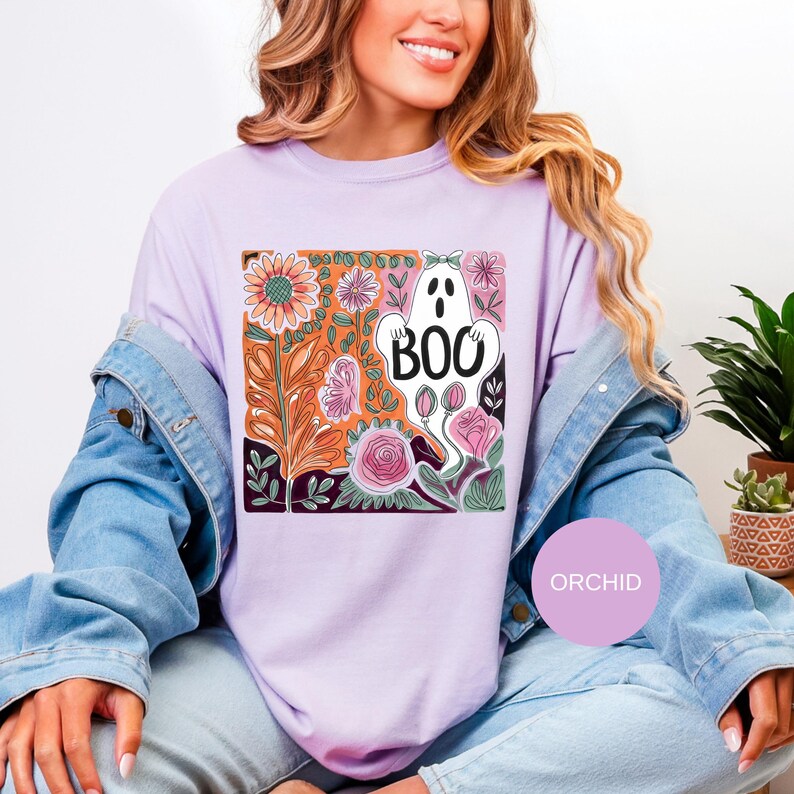 May include: A light purple t-shirt with a colorful graphic featuring a white ghost with the word "BOO" and floral elements. The design includes orange, pink, and green flowers. The shirt is part of a Halloween or fall-themed collection.