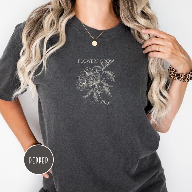 May include: Dark gray t-shirt with a floral design and the text "FLOWERS GROW in the valley" in white. The shirt is a crew neck and has short sleeves. The shirt is made of a soft, comfortable material.