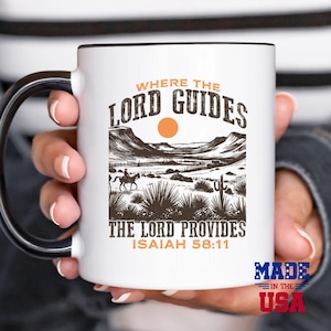 May include: White ceramic mug with a black handle. The mug features a brown and white illustration of a desert landscape with a cowboy riding a horse. The text "Where the Lord Guides, The Lord Provides, Isaiah 58:11" is printed on the mug. The words "Made in the USA" are printed on the bottom of the mug.