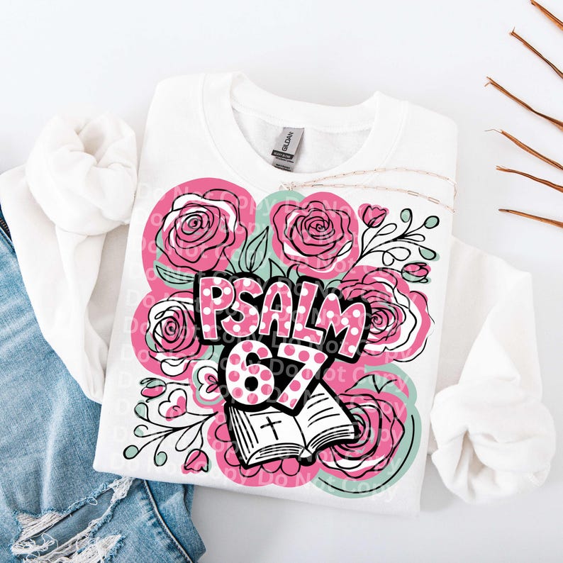 May include: White sweatshirt with a floral design featuring pink roses and the text "PSALM 67." The design includes an open book with a cross. The sweatshirt is folded on top of blue jeans.