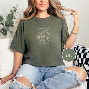 May include: Sage green t-shirt with a floral design and the text "FLOWERS GROW in the valley". The shirt is a relaxed fit and has a crew neck. A round label with the word "SAGE" is visible.
