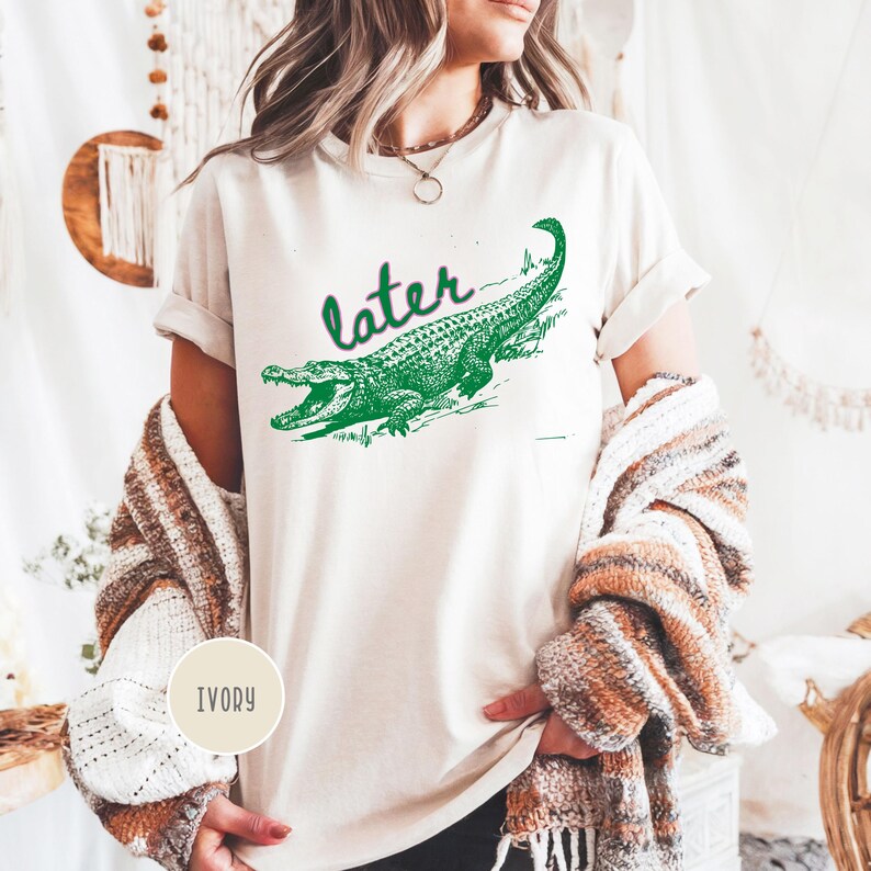 May include: Ivory-colored t-shirt featuring a green alligator graphic with the word "later" above it. The alligator is detailed with scales and open jaws. The shirt has rolled-up sleeves and a casual fit.