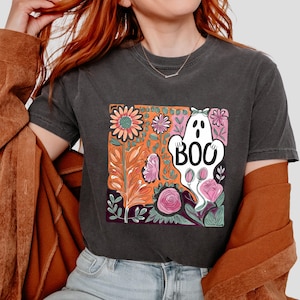 May include: Dark gray t-shirt featuring a colorful floral design with a white ghost that says "BOO". The design includes orange, pink, and purple flowers and foliage, perfect for Halloween or spooky season.