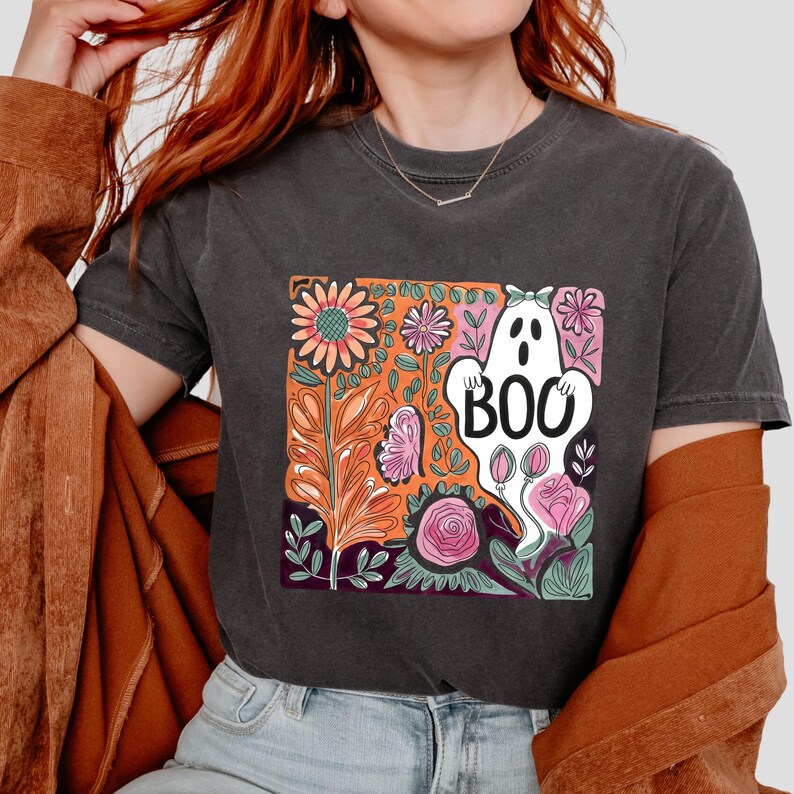May include: Dark gray t-shirt featuring a colorful floral design with a white ghost that says "BOO". The design includes orange, pink, and purple flowers and foliage, perfect for Halloween or spooky season.