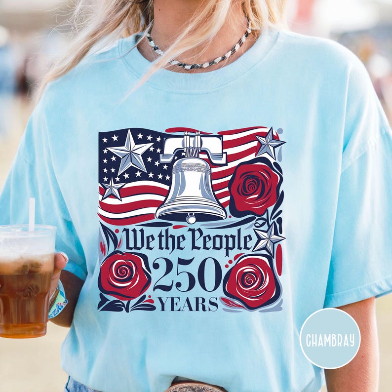 May include: Pale blue t-shirt with a patriotic design. The graphic features the Liberty Bell, American flag elements, red roses, and the text "We the People 250 Years". The shirt has a relaxed fit and short sleeves.