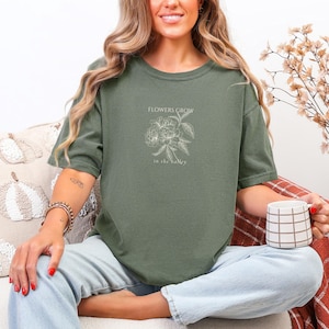 May include: Olive green t-shirt with the text "FLOWERS GROW in the valley" and a floral illustration. The shirt is a relaxed fit and has a crew neck. The person is holding a white and brown checkered mug.