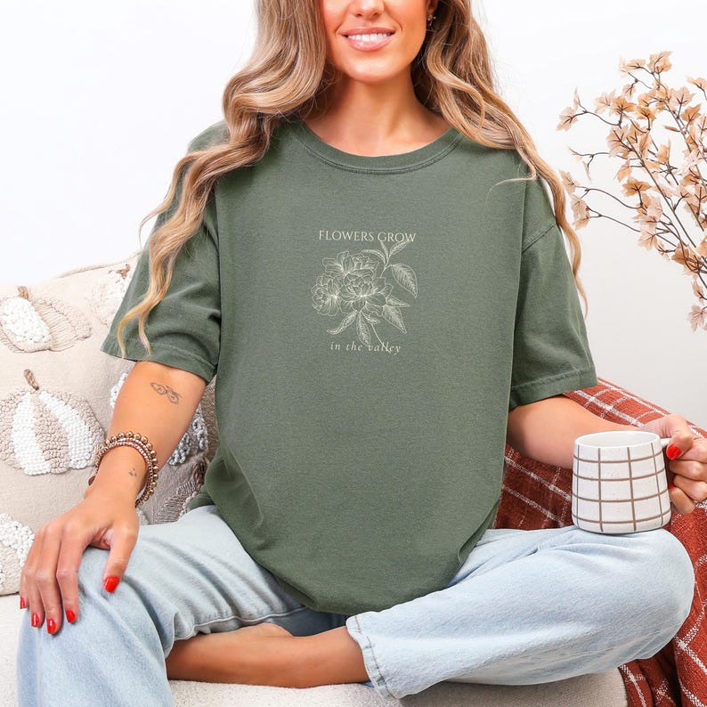 May include: Olive green t-shirt with the text "FLOWERS GROW in the valley" and a floral illustration. The shirt is a relaxed fit and has a crew neck. The person is holding a white and brown checkered mug.