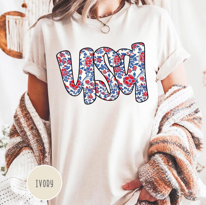 May include: A white t-shirt with the word "USA" printed in red, white, and blue floral print. The shirt is a relaxed fit and has a crew neck. The color of the shirt is ivory.