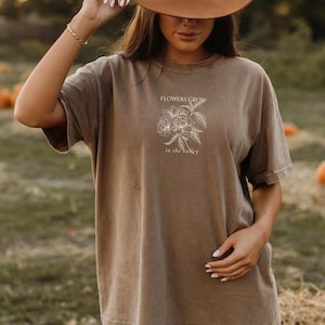 May include: A brown t-shirt with the text "FLOWERS GROW" above a floral illustration and "in the valley" below. The short-sleeved shirt has a relaxed fit and a slightly faded appearance, suggesting a vintage or worn look.