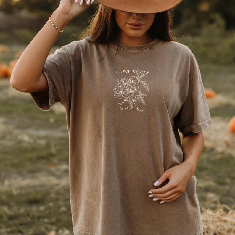 May include: A brown t-shirt with the text "FLOWERS GROW" above a floral illustration and "in the valley" below. The short-sleeved shirt has a relaxed fit and a slightly faded appearance, suggesting a vintage or worn look.