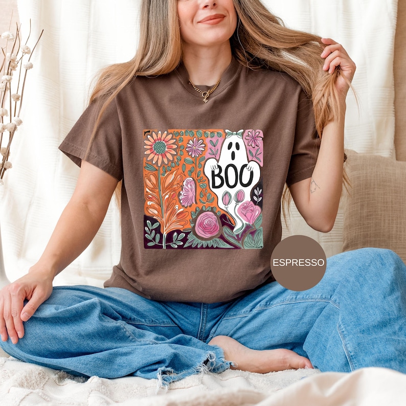 May include: Espresso-colored t-shirt featuring a colorful floral design with a white ghost that says "BOO". The design includes pink roses, orange and purple leaves, and other flowers. The shirt is a comfortable fit.