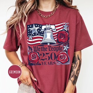 May include: Crimson t-shirt with a patriotic design. The graphic features an American flag, a Liberty Bell, roses, and the text "We the People 250 Years." The design uses red, white, and blue colours.