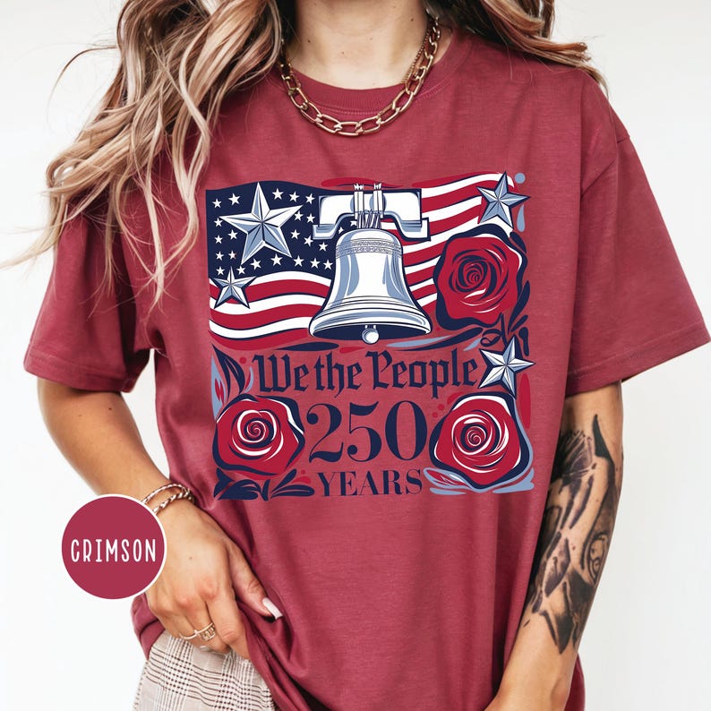 May include: Crimson t-shirt with a patriotic design. The graphic features an American flag, a Liberty Bell, roses, and the text "We the People 250 Years." The design uses red, white, and blue colours.