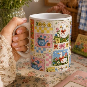 May include: A white ceramic mug with a whimsical garden-themed design. The mug features colorful illustrations of watering cans, ducks, flowers, and the word "SEEDS". The mug is held by a person.
