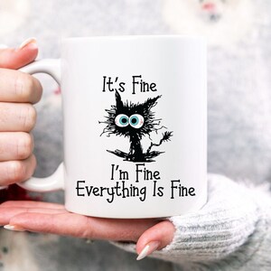 May include: White ceramic mug with a black and white illustration of a cat with the text "It's Fine I'm Fine Everything Is Fine"