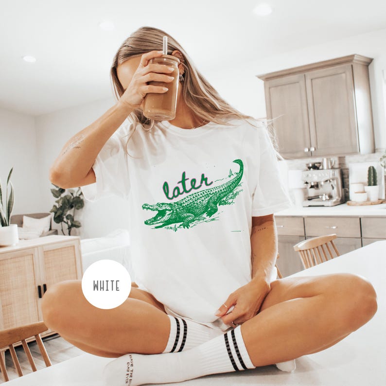 May include: White t-shirt with a green alligator graphic and the word "later" in a script font. The alligator is detailed with scales and open jaws. The shirt is a casual style.