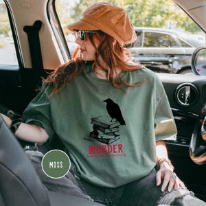 May include: Moss green t-shirt with a black raven perched on a stack of books. The text "MURDER but make it literary" is printed in red below the books. The shirt is a relaxed fit.