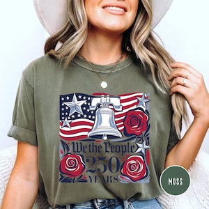 May include: Olive green t-shirt with a patriotic design. The design features the American flag, the Liberty Bell, and red roses, with the text "We the People 250 Years." The shirt is a casual style, suitable for everyday wear.