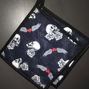 Könnte beinhalten: Zwei schwarze Topflappen mit einem Totenkopf- und Rosenmuster. Die Topflappen haben einen schwarzen Hintergrund mit weißen Totenköpfen, roten Herzen und weißen Rosen. Die Topflappen sind mit schwarzem Stoff eingefasst.