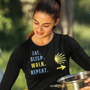May include: A woman wearing a black long-sleeved shirt with the text "Eat. Sleep. Walk. Repeat." and a yellow scallop shell graphic. She is holding a silver pot and a spatula, cooking on a black grill.
