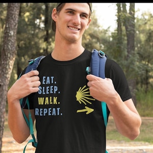 May include: A black t-shirt with the text "Eat. Sleep. Walk. Repeat." and a yellow scallop shell with an arrow pointing to the right. The shirt is being worn by a person in a forest setting.