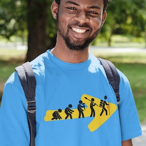 May include: A blue t-shirt with a yellow arrow and a series of stick figures depicting the evolution of humans, ending with a person carrying a backpack and a walking stick. The text "MYCAMINO3D CAMINOWEAR" is visible in the top left corner of the image.