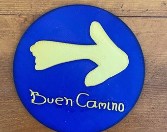 May include: A blue round sign with a yellow arrow pointing to the right. The text "Buen Camino" is written in yellow below the arrow.