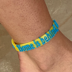 Può includere: Un braccialetto alla caviglia giallo con le parole "Home is behind" in lettere blu. Il braccialetto è indossato sulla caviglia di una persona. Il braccialetto è realizzato in materiale flessibile.