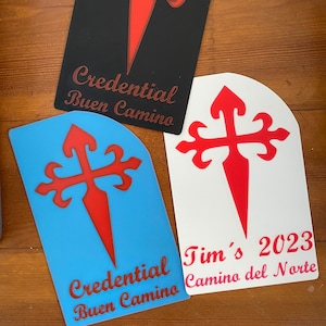 May include: Three pilgrim credential stickers with a red cross and the text "Credential Buen Camino". The sticker on the left is blue, the sticker in the middle is white, and the sticker on the right is black. The white sticker also has the text "Tim's 2023 Camino del Norte".